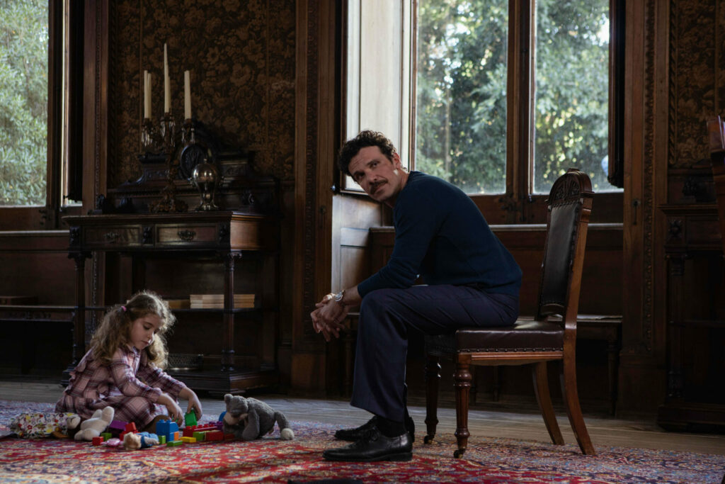Passion of Arts: Saverio Barone sitzt auf einem Holzstuhl, vor ihm auf dem Boden sitzt seine Tochter und spielt auf dem Teppich