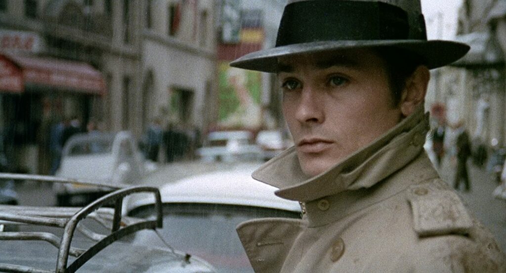 Passion of Arts: Jef Costello steht an seinem Auto und schaut die Straße entlang. Der Antiheld trägt einen Trenchcoat und einen Hut