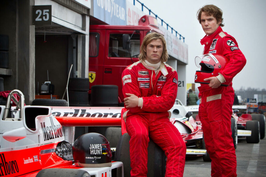 Passion of Arts: Niki Lauda und James Hunt sind bei einem Formel 1 Wagen. Hunt sitzt auf dem Reifen und Niki steht daneben und hält den Helm in der Hand