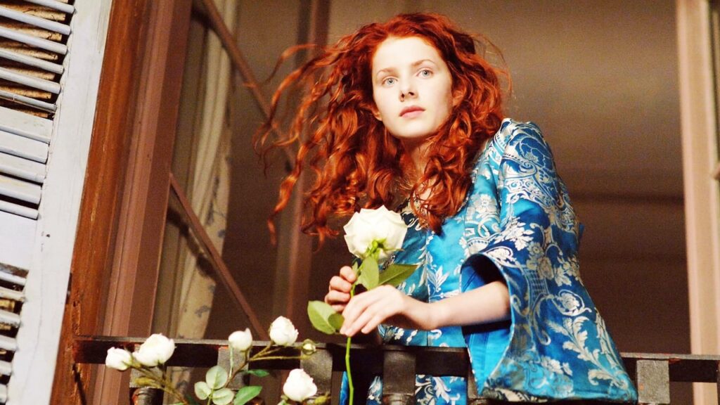 Passion of Arts: Laura steht auf dem Balkon am offenen Fenster. Sie hält eine weiße Rose in der Hand und trägt dabei einen blauen Seidenmantel
