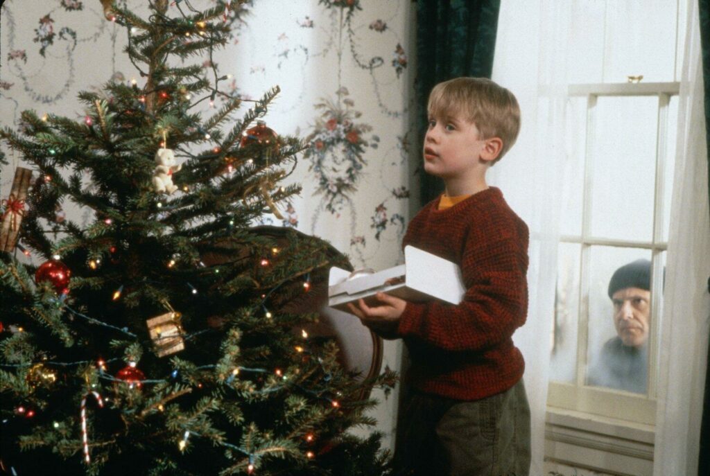 Pasion of Arts Kevin allein zu Haus: Kevin steht vor einem großen Weihnachtsbaum und schmückt ihn. Hinter ihm schaut ein grimmiger Mann zum Fenster rein | Fragen zu Winterfilmen und Weihnachtsfilmen