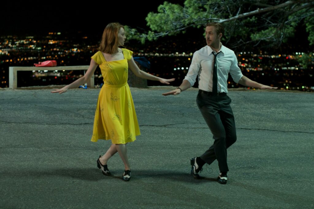 Emma Stone und Ryan Gosling tanzen zusammen. Sie trägt ein gelbes Kleid und er einen Anzug ohne Jackett. Die beiden stehen nebeneinander, haben die Füße überkreuzt und die Arme ausgestreckt. Dabei sehen sie sich an