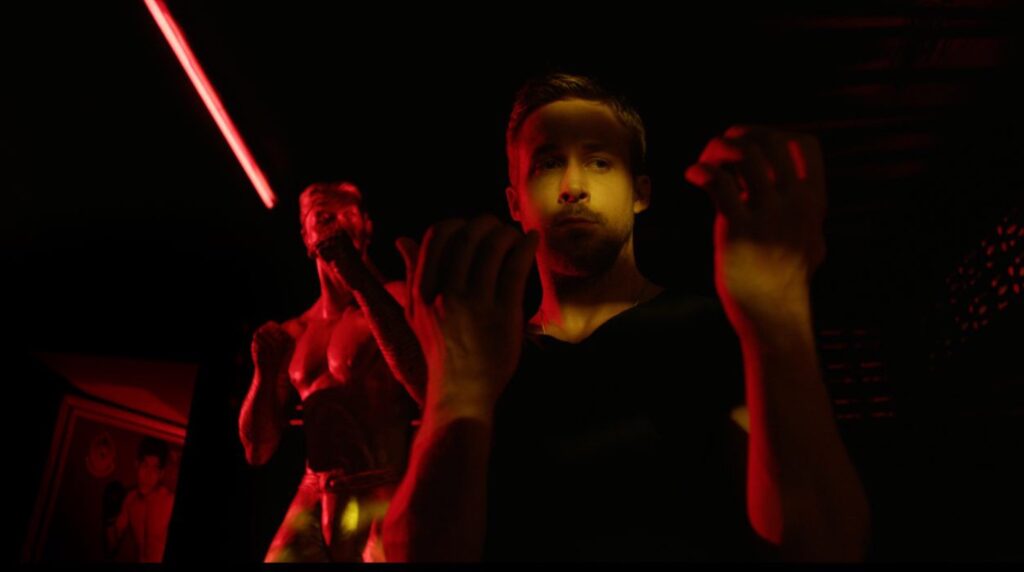 Top 5 Ryan Gosling Performances: Julian steht in einem dunklen Raum, der rot ausgeleuchtet ist. Er hebt die Hände wie zum Boxkampf und starrt darauf. Hinter ihm steht eine Statue von einem Boxer