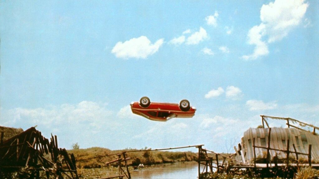 Stunts im Film: Ein rotes Auto schwebt rücklings über den Fluss und springt von einem Holzbrückenteil zum anderen