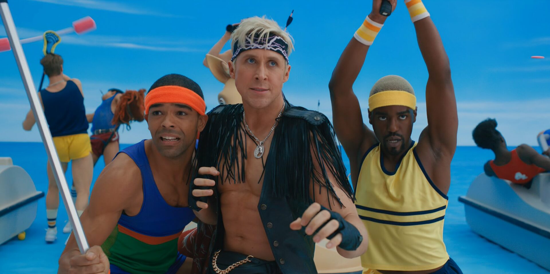 Ryan Gosling ist in der Mitte als Ken im neuen Barbie Film. Er trägt ein Stirnband und eine schwarze Weste mit Fransen. Er ist in Angriffshaltung. Rechts und links neben ihm sind zwei weitere Kens in Kampfhaltung