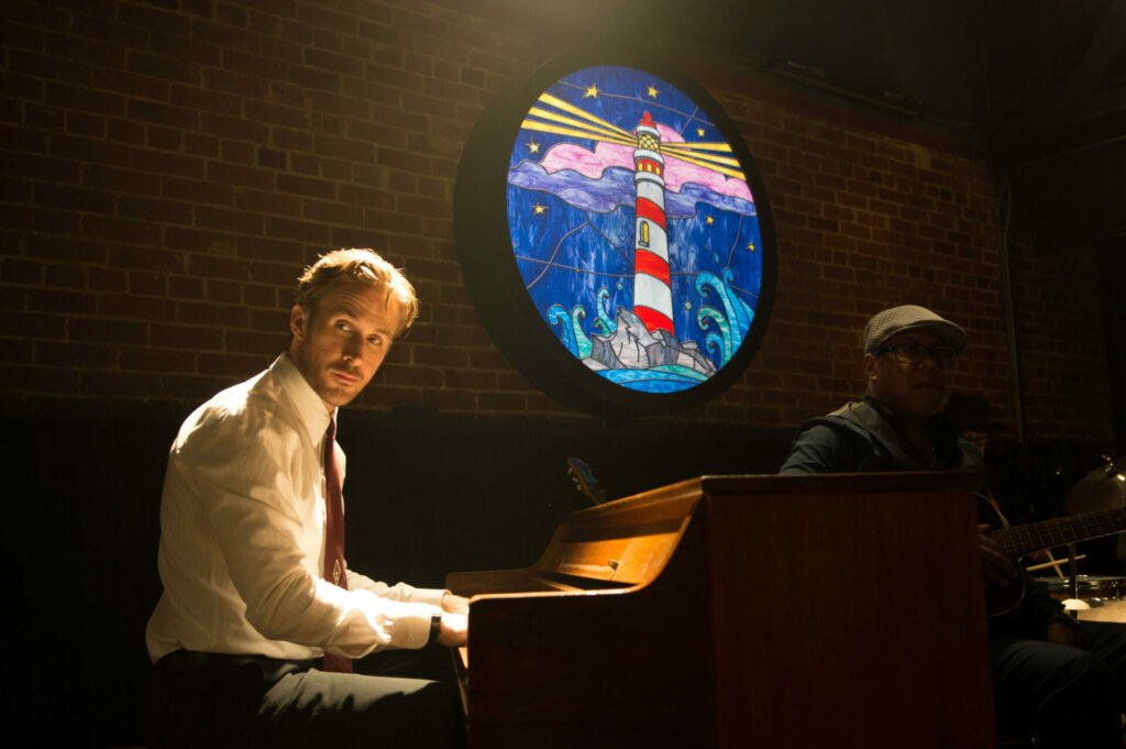 Top 5 Ryan Gosling Performances: Sebastian sitzt am Klavier und schaut zu jemandem im Publikum. Vor seinem Klavier sitzt ein Gitarrist mit Sonnenbrille. Zwischen den beiden hängt oben an der Wand ein gemaltes Bild eines Leuchtturms, das beleuchtet ist