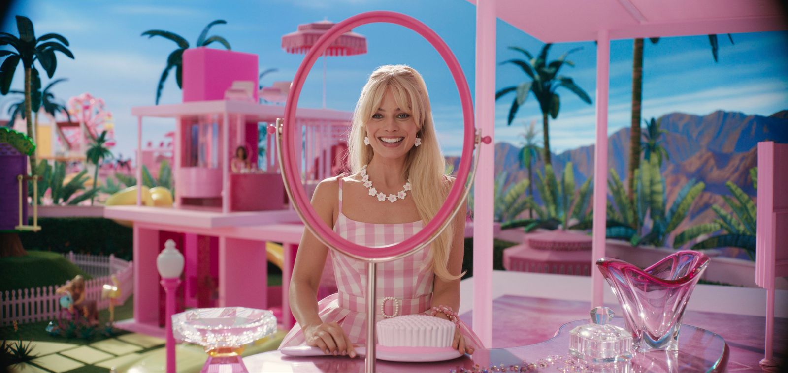 In der Mitte ist Margot Robbie als Barbie und schaut durch einen Spiegel und lächelt. Um sie herum ist alles Pink ebenso ihr Kleid