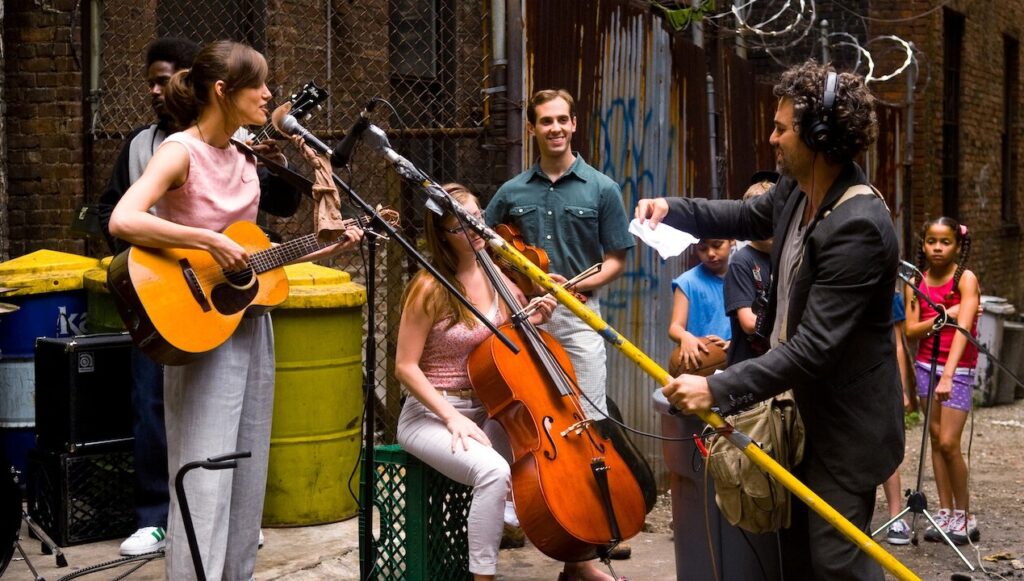 Die 5 besten Sommer in der Stadt Filme: Gretta steht links an der Gitarre und am Mikrofon. Rechts ist Dan, hat Kopfhörer auf, hält ein Mikrofon an einer Stange in der Hand und einen Zettel in der anderen Hand, den er liest. Im Hintergrund ist noch eine Cellistin und ein Geiger, der freundlich lächelt. Daneben stehen Kinder, die zuhören