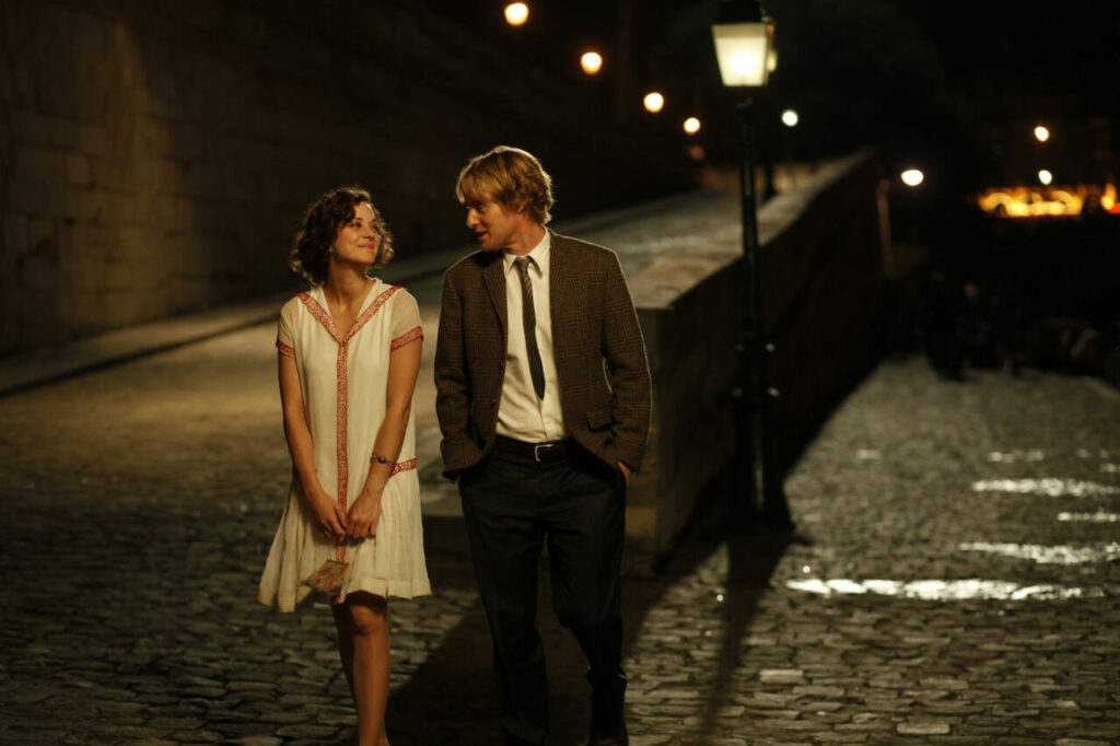 Die 5 besten Sommer in der Stadt Filme: Gil und Adriana gehen nebeneinander spazieren. Sie schauen sich an, während sie gerade eine Straße mit Kopfsteinpflaster passieren. Hinter ihnen ist eine Mauer. Daran entlang befinden sich Straßenlaternen, die im Dunkeln leuchten
