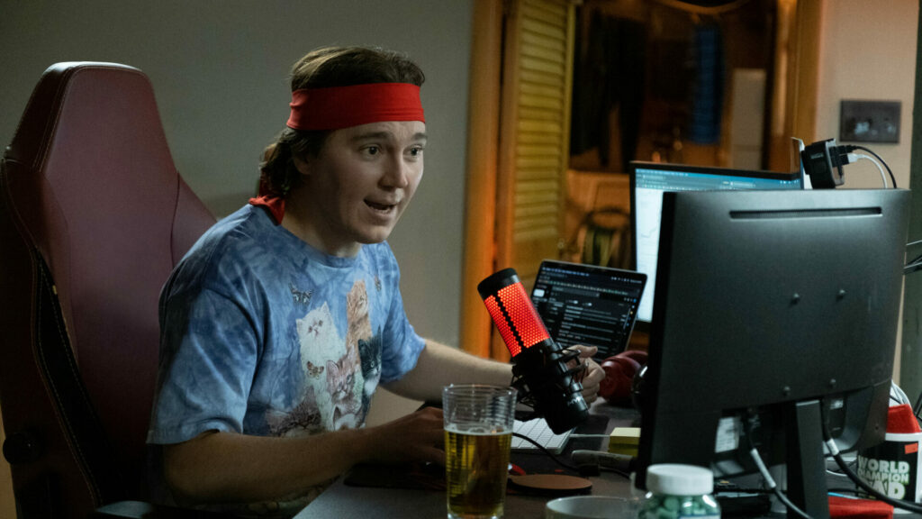 Dumb Money: Paul Dano als Keith Gill. Er sitzt an einem Schreibtisch. Vor ihm sind zwei Bildschirme und ein rotes Mikrofon. Neben ihm steht ein Bierglas, er trägt ein Katzen T-Shirt und ein rotes Stirnband
