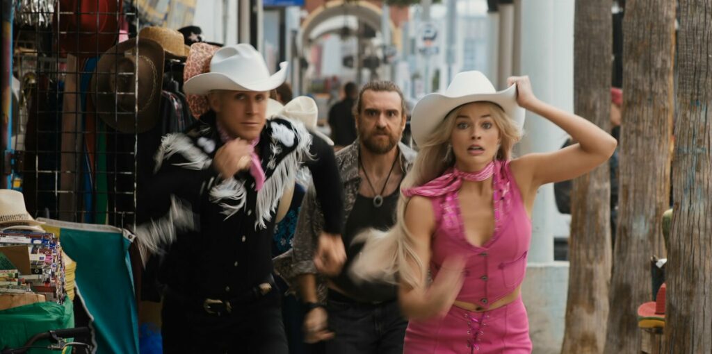 Ken und Barbie tragen Cowboykostüme und rennen vor einem bärtigen Mann davon. Barbie hält dabei ihren Hut fest