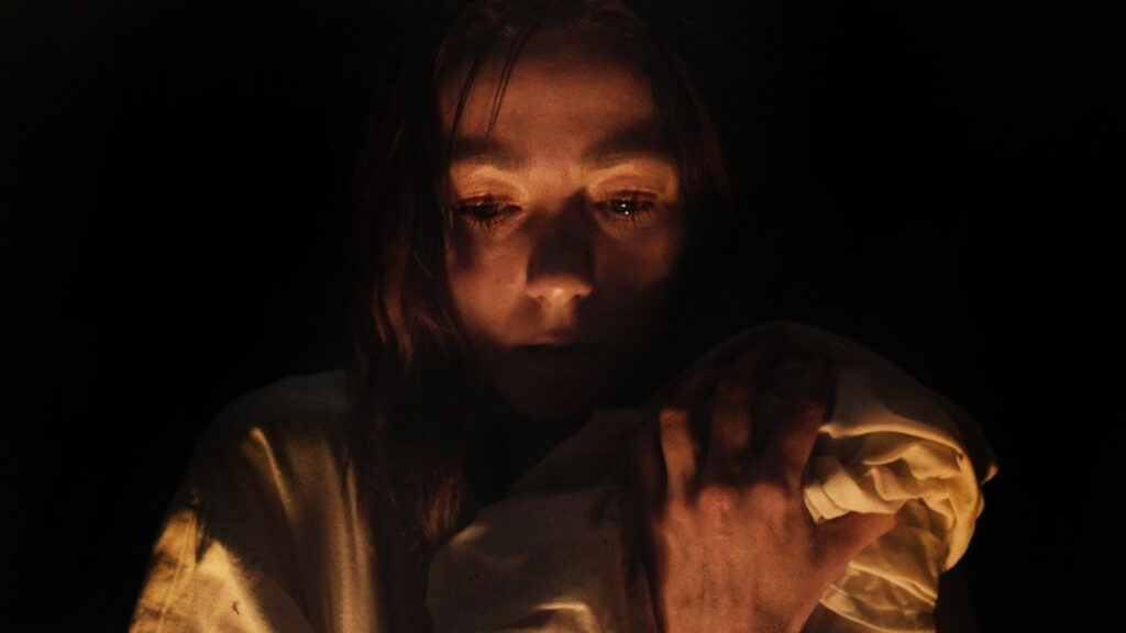 Hagazussa - Der Hexenfluch: Man sieht Albrun (Aleksandra Cwen) von vorne. Um sie herum ist alles dunkel. Ein schwacher Kerzenschein erhellt ihr Gesicht. Sie hat Tränen in den Augen und hält ein Bündel ganz fest im Arm an ihre Schulter