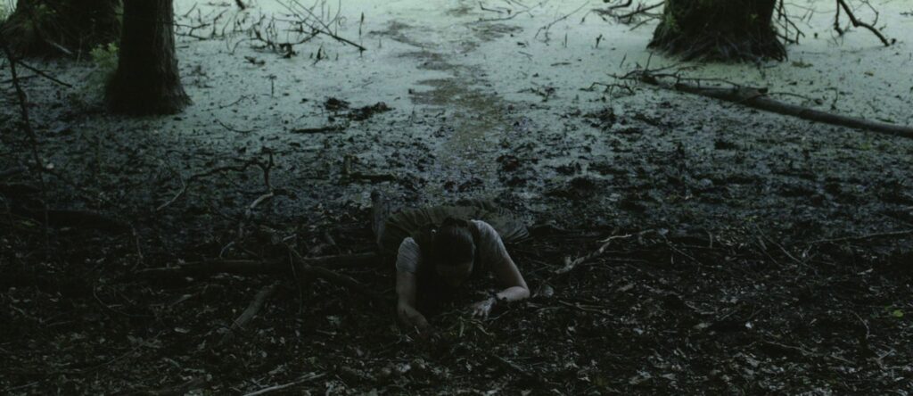 Albrun (Aleksandra Cwen) liegt im Schlamm vor dem See und krabbelt aus dem Wasser heraus