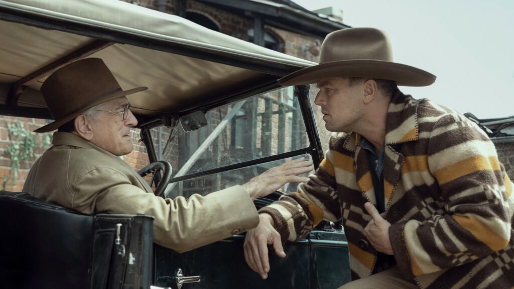 Links ist William Hale in einem Auto. Er trägt einen braunen Hut und eine Brille. Er redet auf Ernest ein, der neben ihm an der Autotür steht. Er trägt einen Cowboyhut und eine bunt gestreifte Jacke