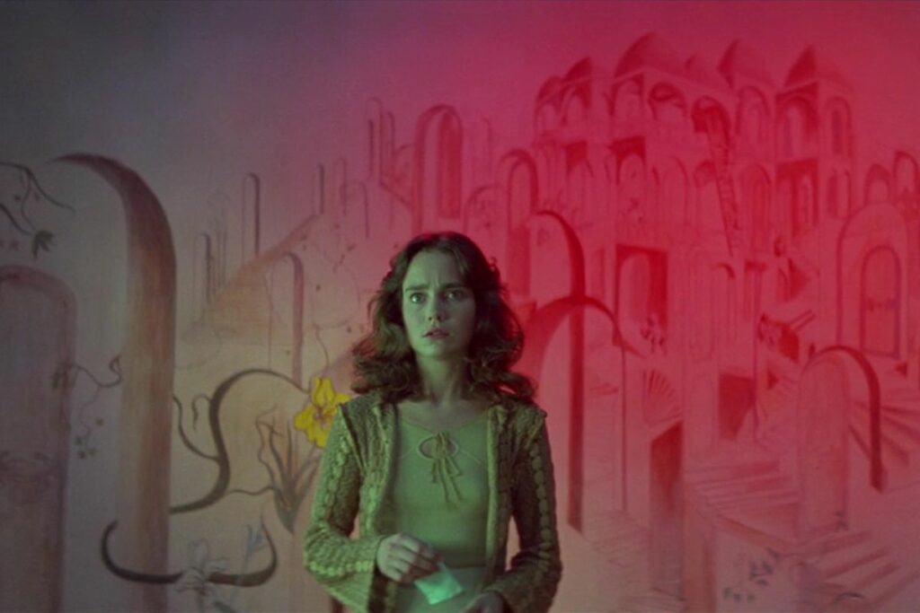 Media Monday #643: Suzy steht vor einer rot beleuchteten Wand, die mit Rundbögen und Treppen bemalt ist. Ihr Gelbgrünes Gewand bietet einen Kontrast zum Hintergrund. Sie hält einen Notizzettel in der Hand und schaut ängstlich