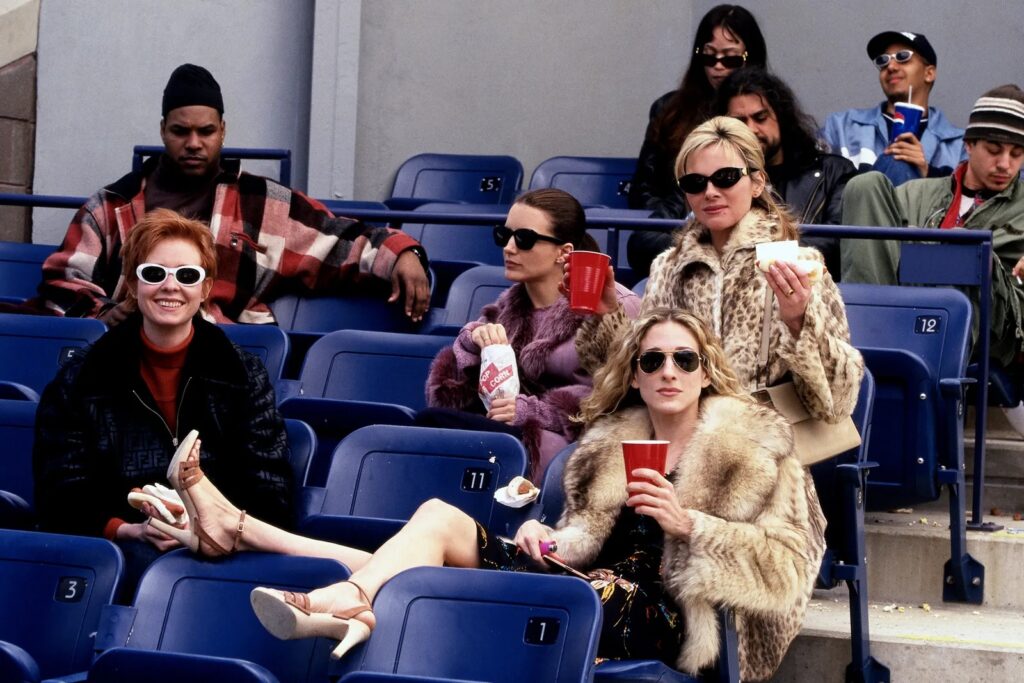 Auf dem Bild sieht man vier Frauen, die Sonnenbrillen tragen und es sich auf der Tribüne eines Stadions bequem gemacht haben. Sie haben Pappbecher mit Bier in der Hand und Hot Dogs. In den hinteren Reihen sind noch ein paar andere Zuschauer*innen