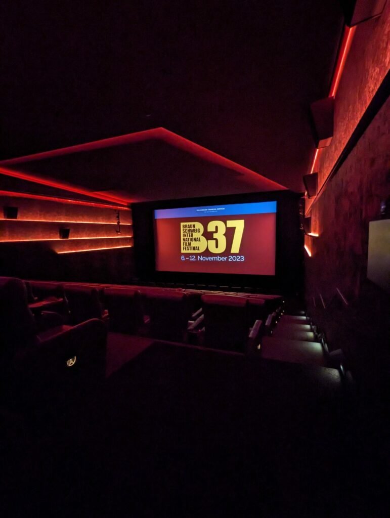 Braunschweig International Film Festival: Das Bild zeigt einen roten Kinosaal der nur von der Leinwand beleuchtet ist, auf der das Logo des Festivals zu sehen ist