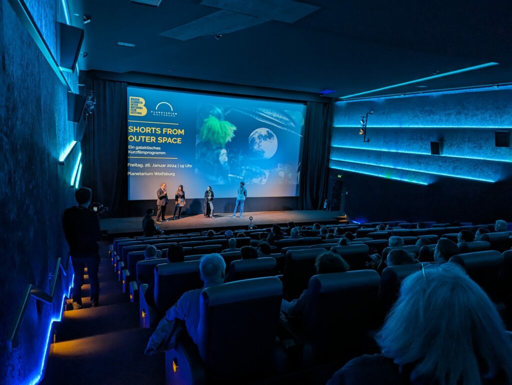 Braunschweig International Film Festival. Man sieht einen blau beleuchteten Kinosaal. Vorne stehen Menschen. Ein Mann spricht in ein Mikrophon.
