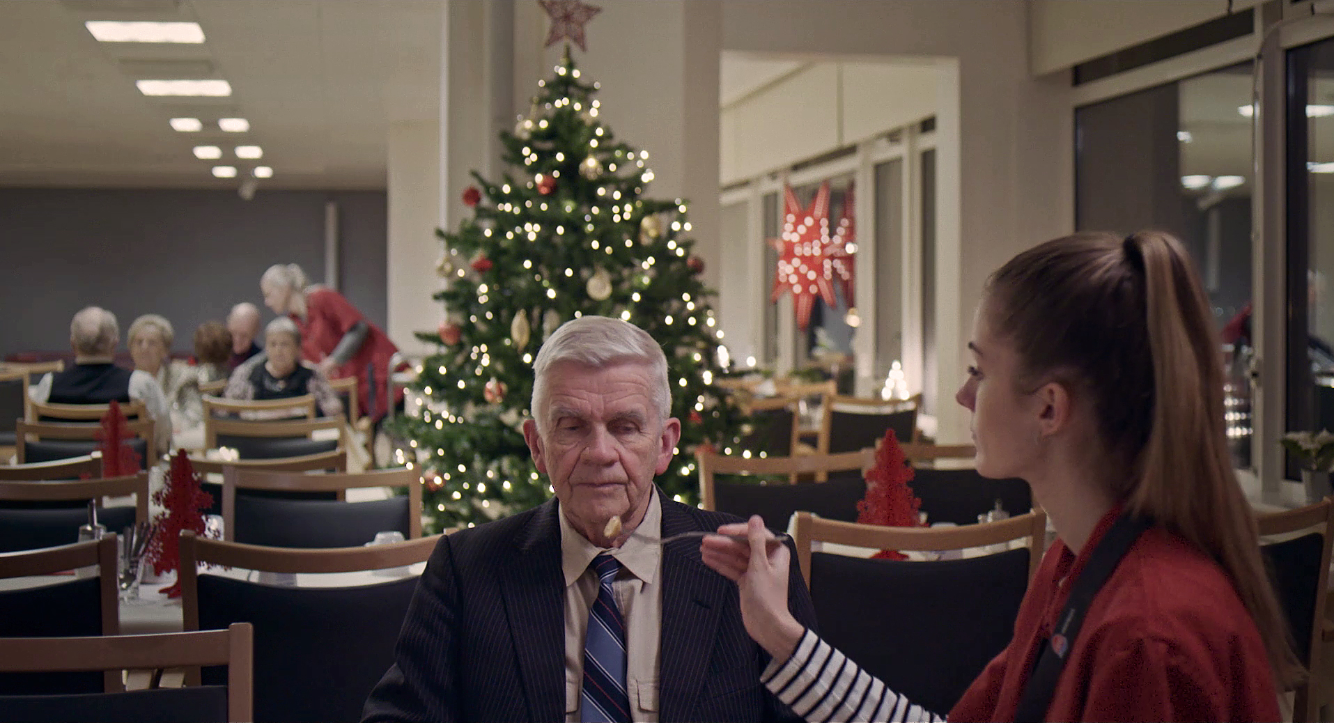 Echo: Auf dem Bild ist ein Mann im Anzug, der von einer jüngeren Frau gefüttert wird. Im Hintergrund steht ein geschmückter Weihnachtsbaum, viele Stühle und mehrere ältere Menschen sitzen an Tischen