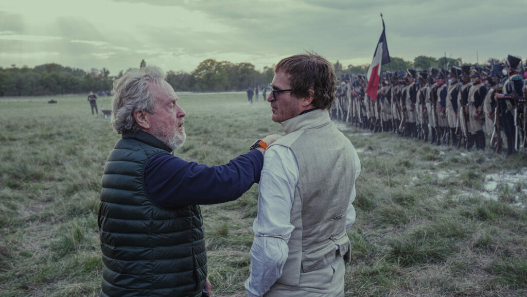 Auf dem Bild sind links Ridley Scott, der den Kragen von Joaquin Phoenix richtet, der rechts im Bild steht und eine Sonnenbrille trägt. Ganz rechts im Bild sind Statisten im Soldatenkostüm in einer Reihe