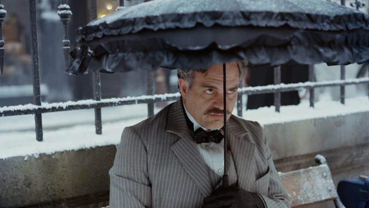 Auf welchen Film im Jahr 2024 freust du dich schon besonders? Auf dem Bild ist ein Mann zu sehen, der auf einer Parkbank sitzt. Hinter ihm ist ein Gitter zu sehen, das Schnee bedeckt ist. Er hält den Kopf gesenkt, aber den Blick nach oben gerichtet. Er schützt sich vor dem nassen Wetter mit einem großen, blauen Schirm