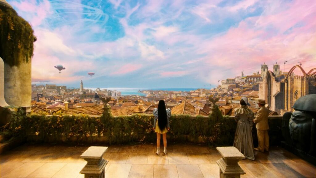 Auf dem Bild ist eine Frau und ein Pärchen zu sehen, das auf einer Terrasse steht. Sie blicken auf eine bunte Stadt herunter