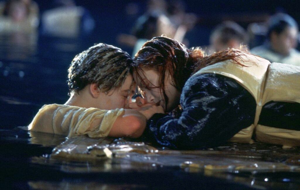 Top 5 emotionale Liebesfilme, die zum Weinen bringen: Auf dem Bild sieht man ein Paar, das im Eiswasser ist. Sie liegt auf einer Holztür und trägt eine Rettungsweste. Er ist vor ihr im Wasser und hält liebevoll ihre Hände