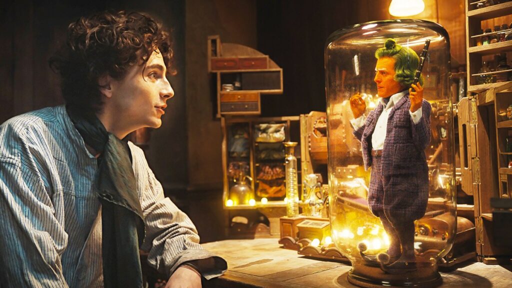 Auf dem Bild sieht man einen kleinen Mann mit oranger Haut und grünen Haaren, der auf dem Tisch unter einem Glas steht. Auf der linken Seite sitzt Willy Wonka und spricht mit dem Mann