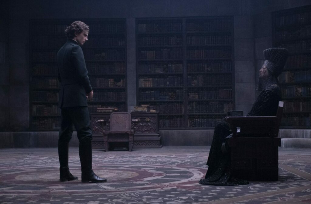 Auf dem Bild sieht man zwei Menschen in einer Bibliothek. Im Hintergrund sind viele Regale gefüllt mit Büchern. Eine Person sitzt auf einem prunkvollen Holzstuhl, in schwarz gekleidet und verschleiert. Die andere Person steht im schwarzen Anzug vor der sitzenden Person und hat den Kopf gesenkt