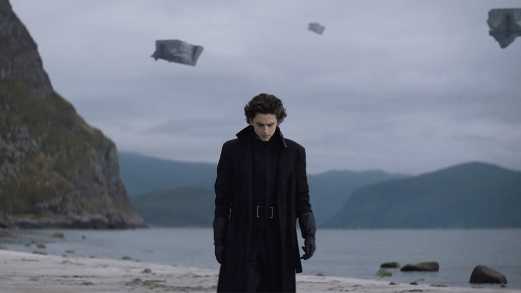 Auf dem Bild ist ein junger Mann, der den Blick gesenkt hat. Er trägt schwarze Kleidung, einen langen Mantel, Lederhandschuhe und einen Gürtel mit einer großen Schnalle. Im Hintergrund sind Berge zu sehen und das Meer. Außerdem schweben im Himmel irgendwelche Gegenstände