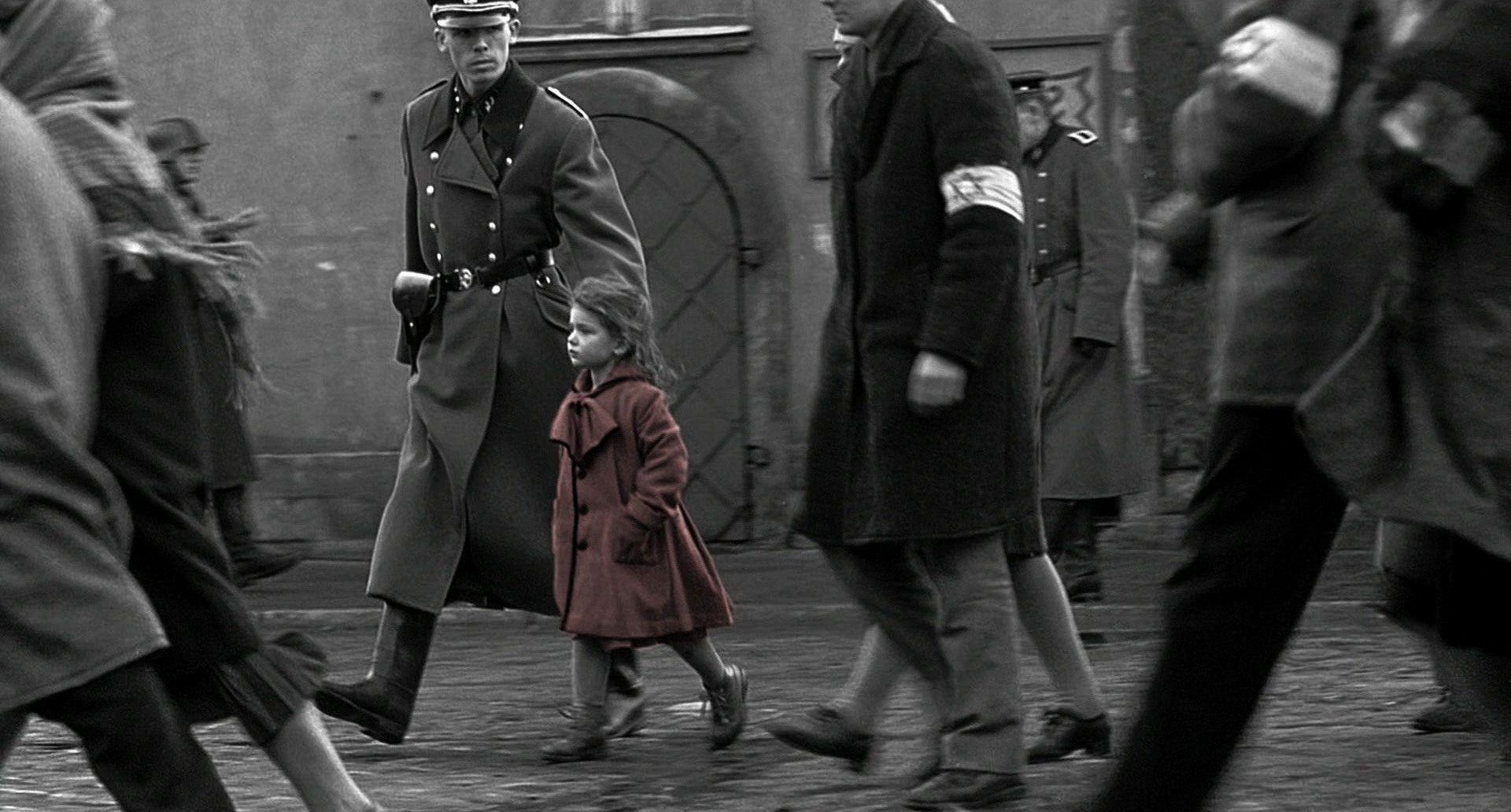 Kameraarbeit: Auf dem Bild sieht man einen SS Soldaten, der neben einem kleinen Mädchen im roten Mantel geht. Um die beiden herum gehen noch ein paar weitere Menschen mit Armbinden, die den Davidstern zeigen