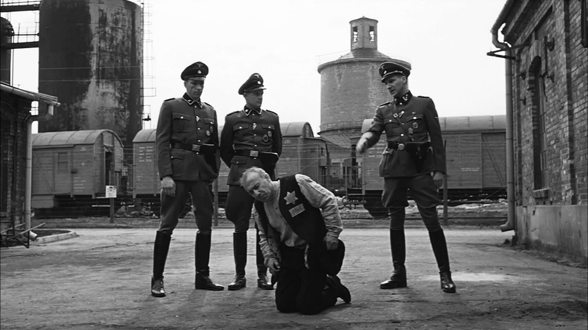 Kameraarbeit: Auf dem Bild sind drei SS Soldaten zu sehen, die um einen knieenden Mann stehen. Im Hintergrund ist ein Fabrikgebäude zu sehen. Davor ist ein Zug.