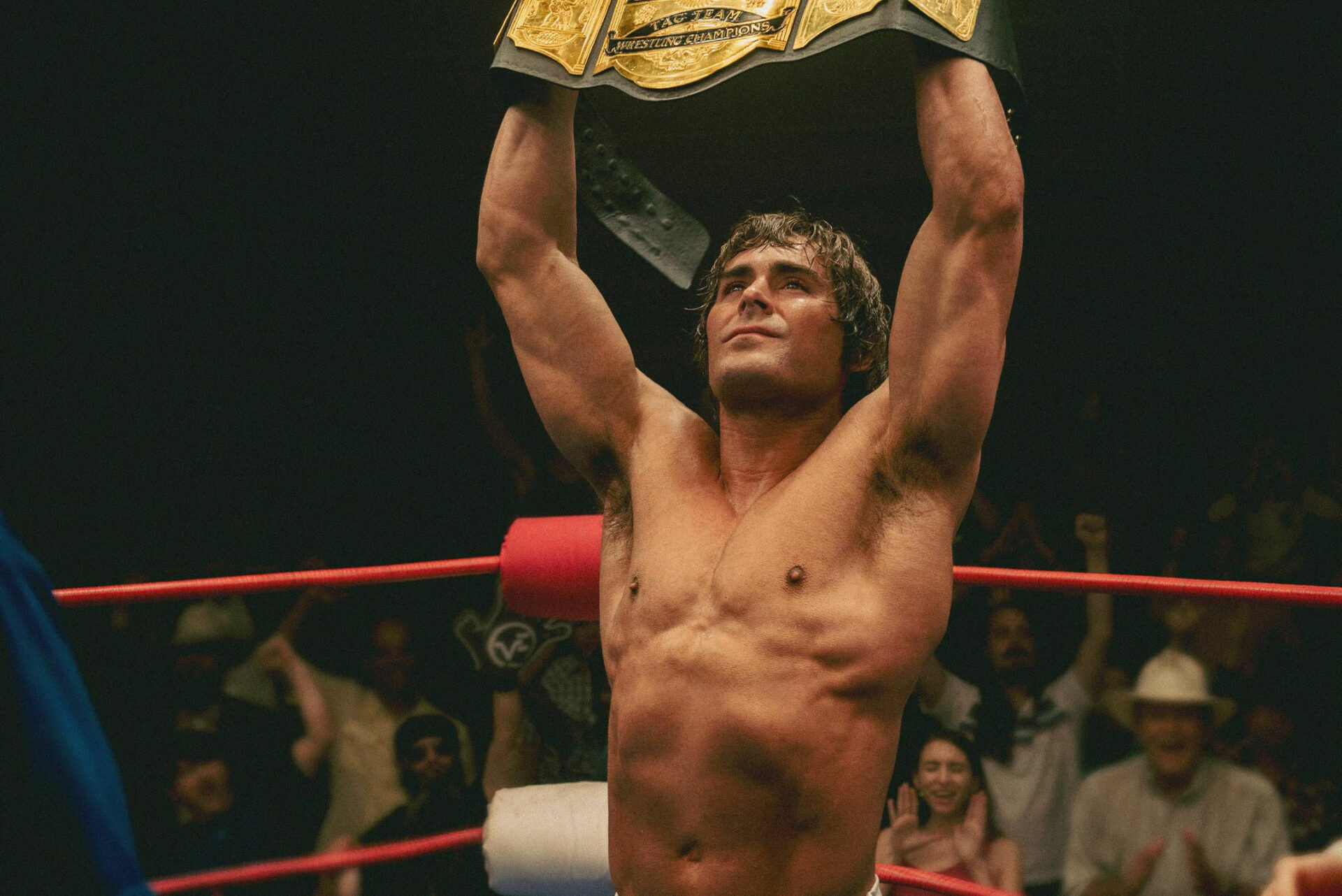 Das Bild zeigt einen jungen Mann, der den Oberkörper frei hat, in einem Boxring steht und einen goldenen Gürtel in die Höhe hält. Hinter ihm sieht man ein jubelndes Publikum