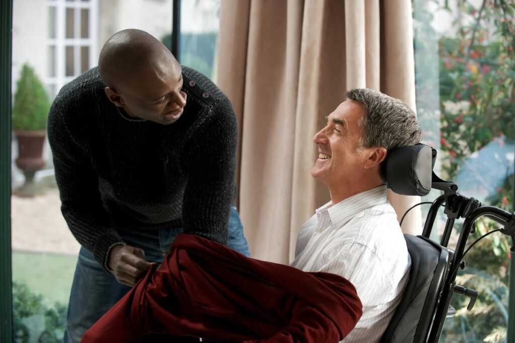 Fragen zu Buchverfilmungen: Das Bild zeigt zwei Männer. Einer sitzt im Rollstuhl und wird von dem anderen gerade zugedeckt. Beide Lachen herzhaft