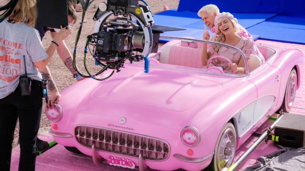 Top Filme mit atemberaubenden visuellen Effekten: Auf dem Bild ist ein rosa Auto zu sehen, in dem ein Mann und eine Frau sitzen. Beide lachen. Vor dem Auto hängt eine Kamera zum Filmen und dahinter sind Menschen vom Set zu sehen