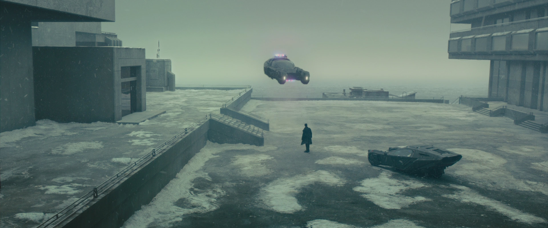 Blade Runner 2049: Das Bild zeigt einen Mann, der vor einem großen Gebäude steht. Um ihn herum ist alles vereist und verschneit. Hinter ihm steht ein futuristisches Fahrzeug. Vor ihm ist ein Polizeiauto, das ebenfalls futuristisch ist und in der Luft schwebt