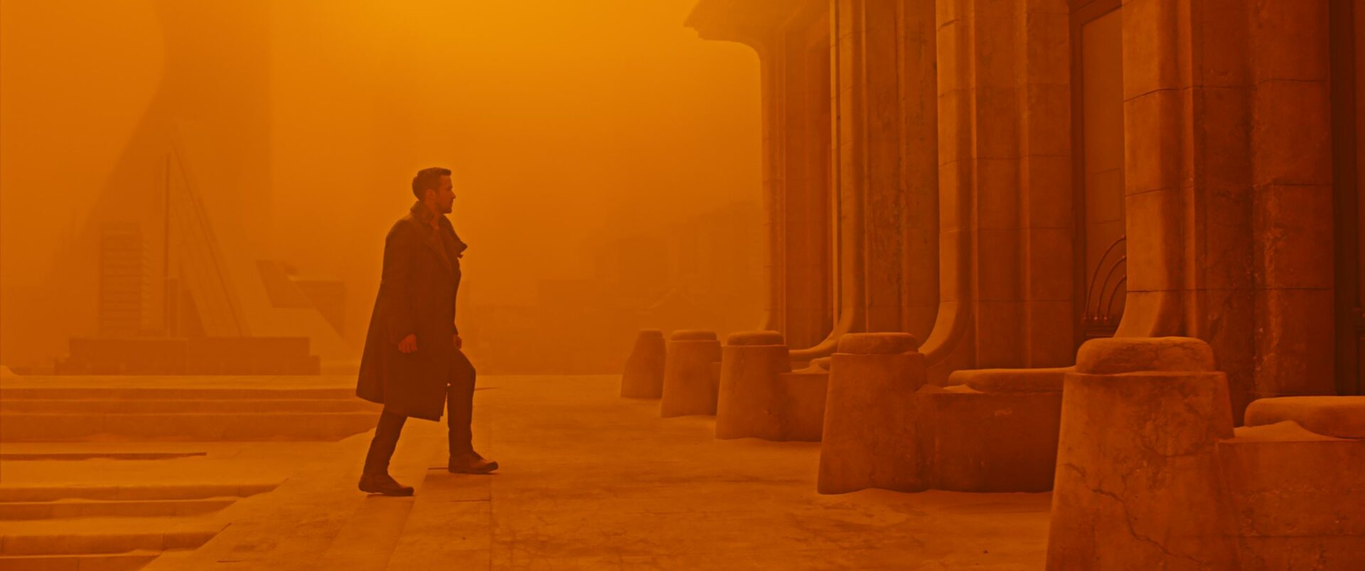 Blade Runner 2049: Auf dem Bild ist ein Mann zu sehen, der gerade ein paar Treppen hinauf zu einem Gebäude geht. Das Ganze Bild ist in gelb orange Tönen