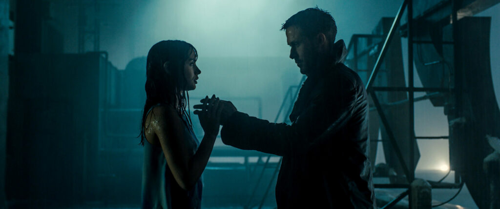 Blade Runner 2049: Das Bild zeigt einen Mann und eine Frau, die sich im dunklen, blauen Licht gegenüber stehen. Sie halten sich an den Händen. Im Hintergrund sind Maschinen zu sehen