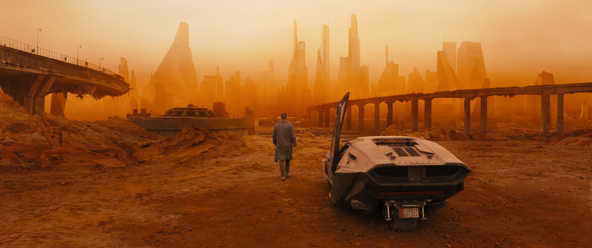 Blade Runner 2049: Das Bild zeigt einen Mann, der gerade aus einem futuristischen Auto gestiegen ist. Die Tür davon steht noch offen. Er geht auf eine tote Stadt zu, die in einem orangen Nebel verschwimmt