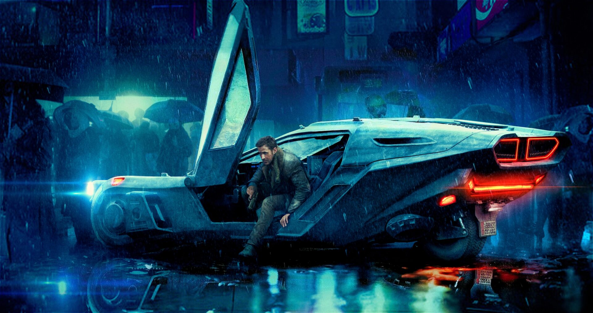 Blade Runner 2049: Das Bild zeigt einen Mann, der gerade aus einem futuristischen Auto steigt. Im Hintergrund sind Gebäude zu sehen. Davor ist eine Menschenmenge, die sich mit Schirmen vor dem Regen schützt