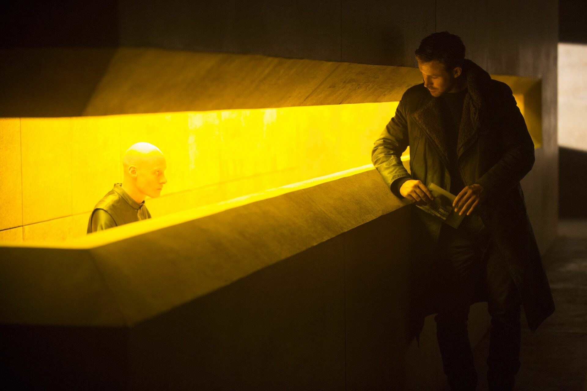 Blade Runner 2049: Das Bild zeigt einen Mann, der an eine Art Empfangstresen lehnt. Am Tresen sitzt ein glatzköpfiger Mann, der zu dem anderen hinauf sieht. Der Tresen ist aus Beton und zeigt nur einen kleinen Schlitz der gelb leuchtet, aus dem der glatzköpfige Mann heraus blickt