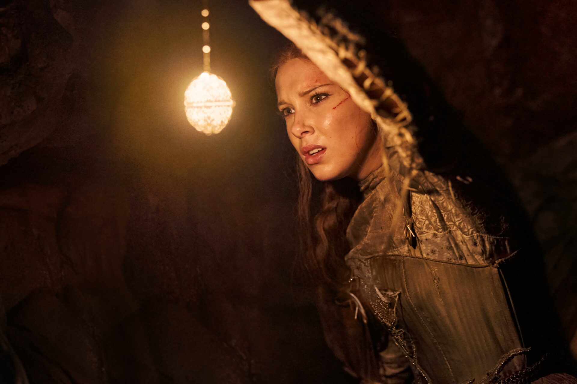 Damsel: Das Bild zeigt eine junge Frau, die in einer dunklen Höhle ist und mit einer Art Medaillon den Weg leuchtet.
