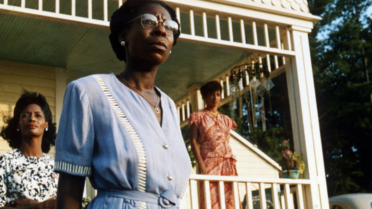 Die Farbe Lila: Das Bild zeigt drei Frauen, Eine steht hinten auf der Veranda, eine davor, etwas daneben. Im Vordergrund ist eine Frau mit Brille und blauem Kleid. Sie schaut etwas skeptisch