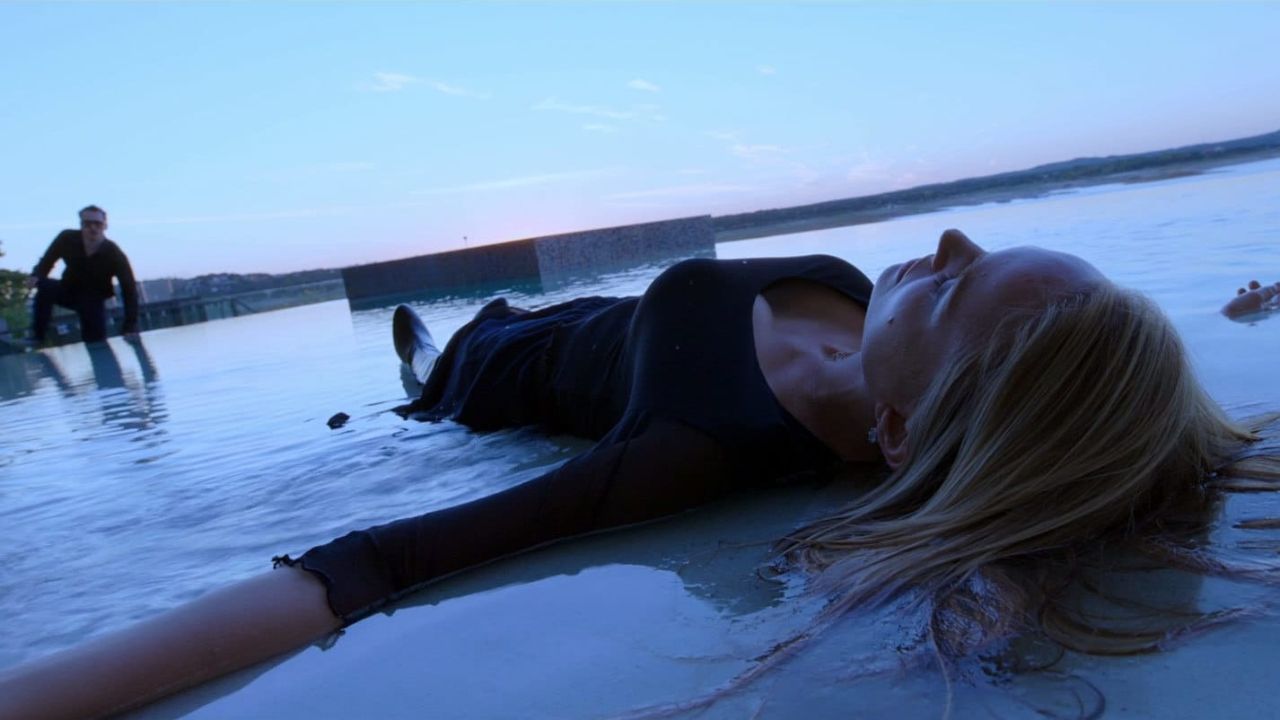 Das Bild zeigt eine Frau, die am Strand liegt, die Augen geschlossen und die Arme ausgebreitet, liegt sie im Wasser mit dem Gesicht zum Himmel. Im Hintergrund bewegt sich ein Mann auf sie zu