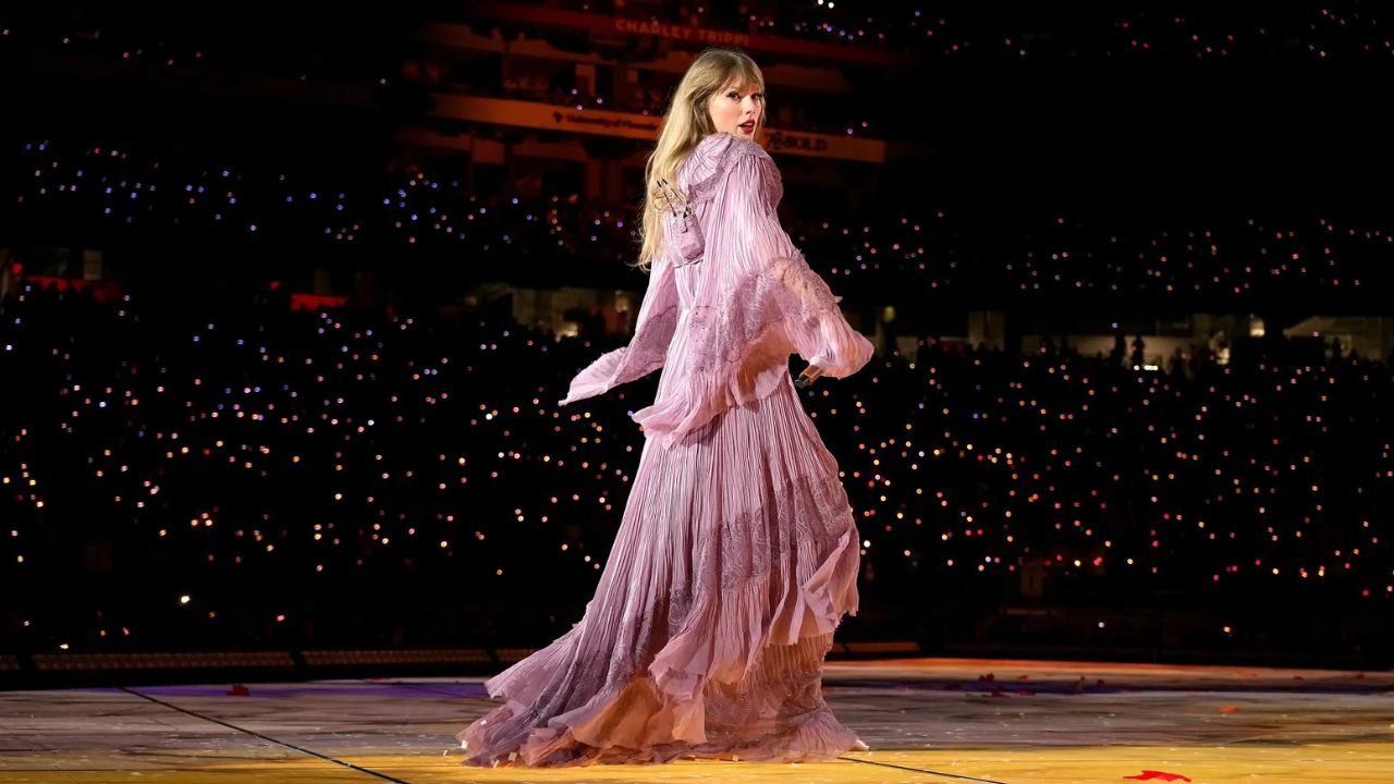 Das Bild zeigt Taylor Swift auf der Bühne in einem flatternden lilafarbenen Gewand. Sie tanzt vor einem Publikum, das sich im Hintergrund nur durch Handylicher bemerkbar macht