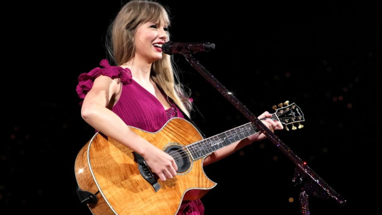 Taylor Swift: The Eras Tour: Auf dem Bild hält Taylor Swift eine Akustik Gitarre in der Hand und spielt darauf. Vor ihr ist ein Mikrofon. Sie lächelt