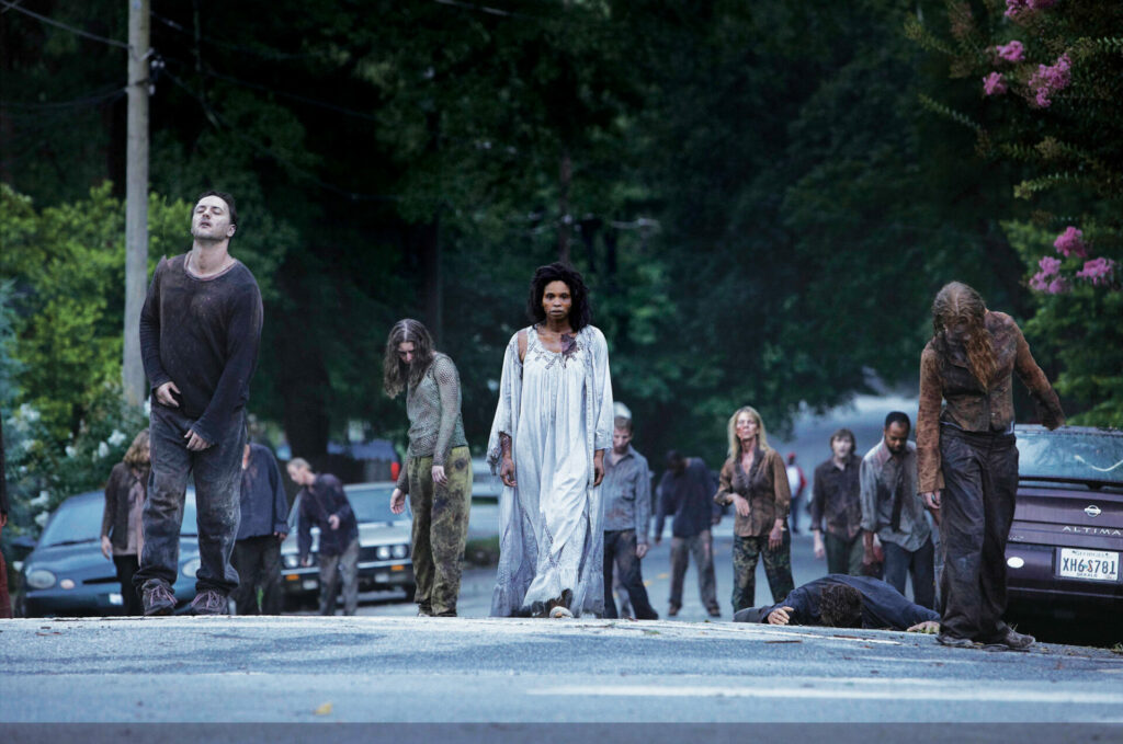 Auf dem Bild sind Zombies, die die Straße entlang gehen. In der Mitte ist eine Frau in einem weißen Gewand
