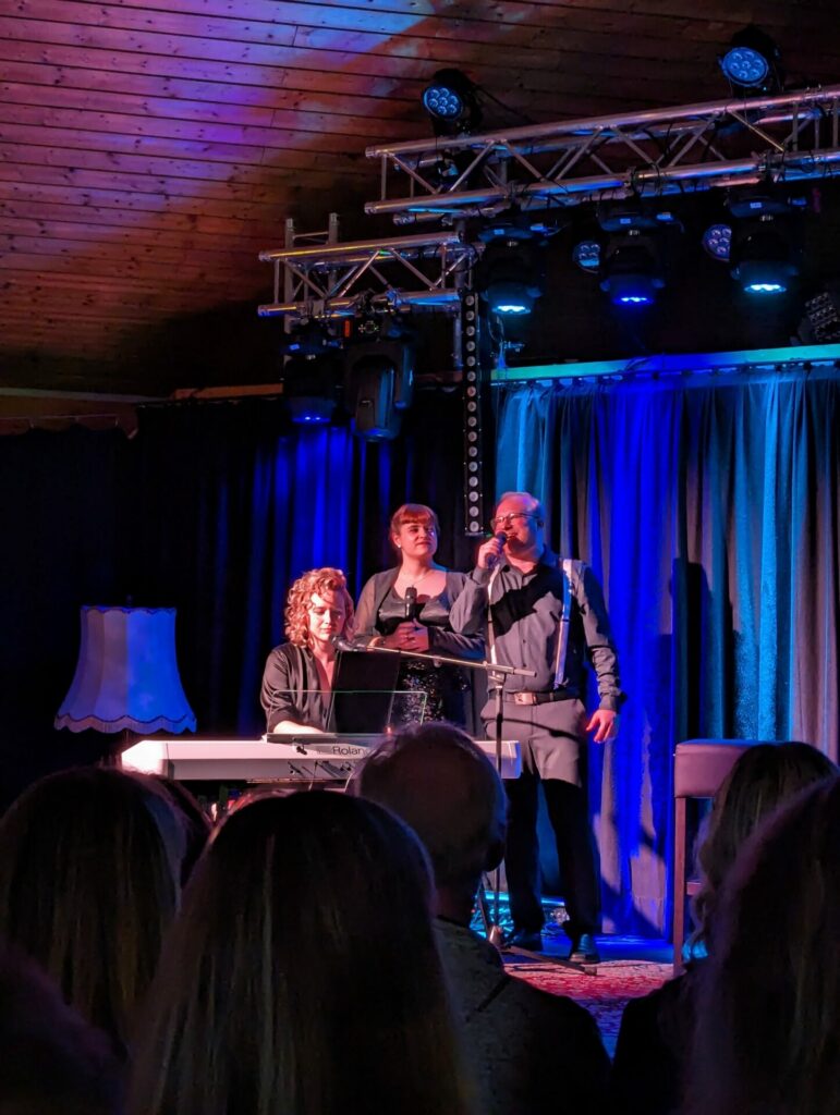 Passivattraktiv: Auf dem Bild ist eine Musikgruppe. Eine Frau sitzt am Klavier, eine Frau steht mit Mikrofon in der Mitte und ein Mann spricht gerade ins Mikrofon