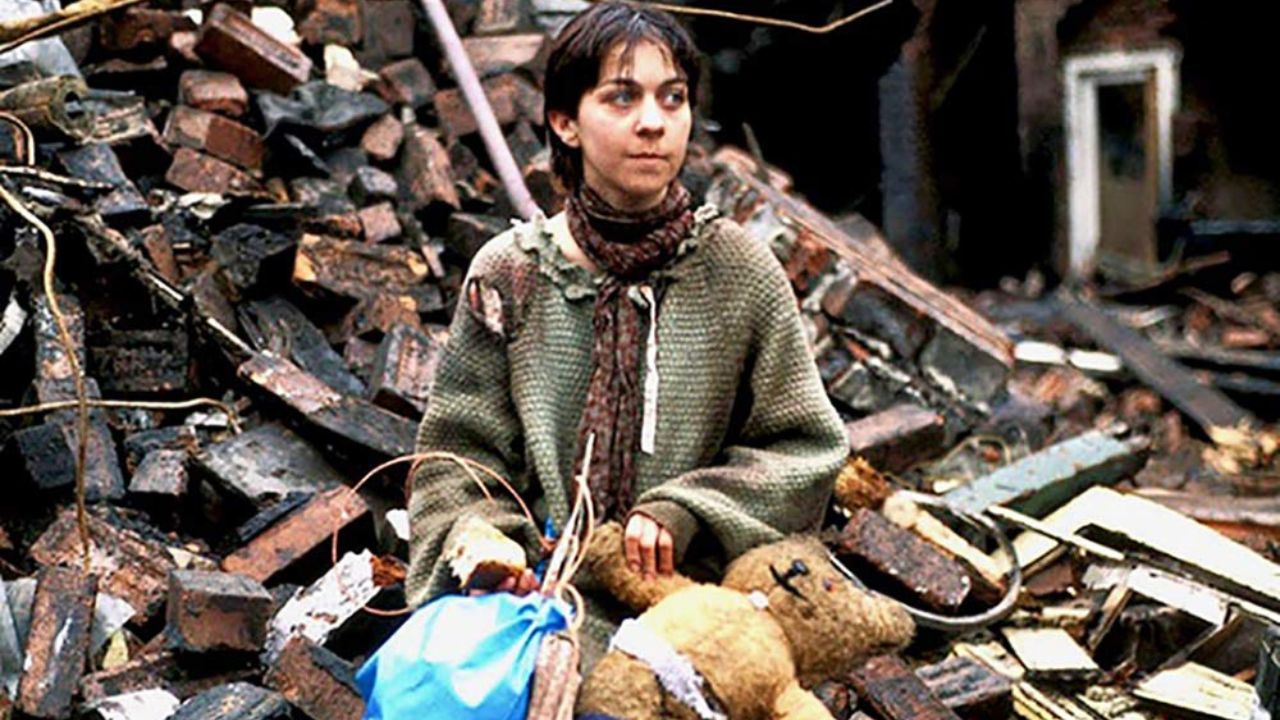 Threads: Das Bild zeigt eine junge Frau, die zerfetzte Kleidung trägt und in einem Trümmerhaufen sitzt. Vor ihr auf dem Schoß hat sie einen Teddybären