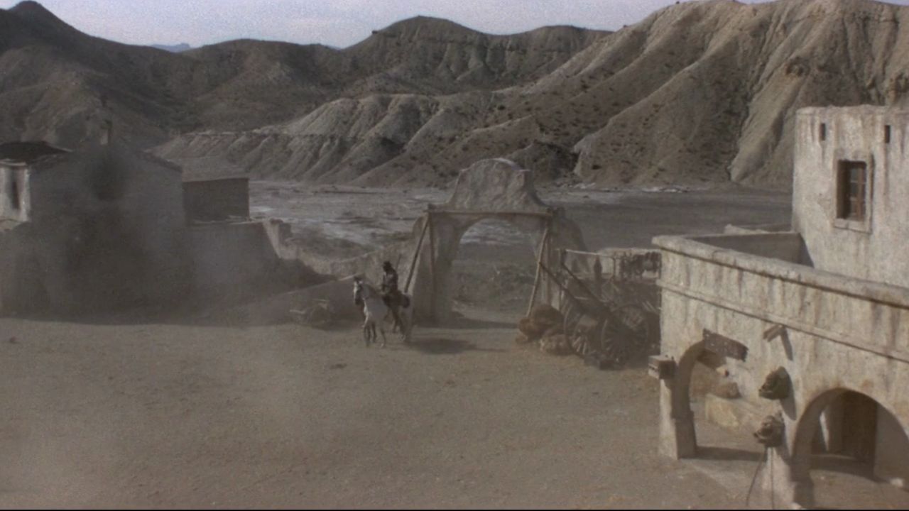 Auf dem Bild sieht man eine Wüstenlandschaft. Auf der rechten Seite ist ein weißes Gebäude, in der Mitte reitet gerade jemand zu einem Tor herein
